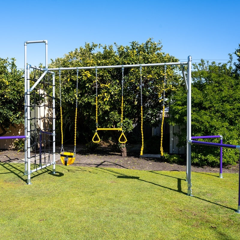 A Funky Monkey Bars play set stands on a tidy lawn, featuring a cargo net, toddler swing, gym rings, trapeze bar, and bold purple parallel bars. Bright yellow chains and a backdrop of green trees and blue sky create a fun outdoor space.