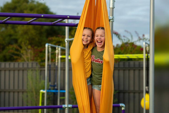 Two children sitting in orange Aerial Silk on a Funky Monkey Bars frame