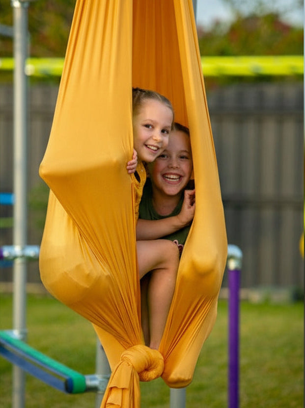 Child and adult peeking through orange Aerial Silk on a backyard frame